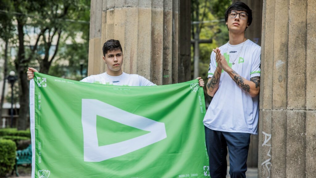 LOUD midlaner TinOwns rubs his hands together while LOUD AD Carry Brance holds a flag bearing the LOUD logo of a white right-facing triangle on a field of green. Both players look out of shot to the right.