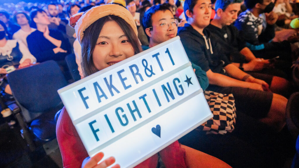 Image shows an Asian woman wearing a soft hat with ears holding a lightbox to the camera that reads Faker & T1 fighting followed by a star and a heart
